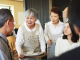 The Restaurant of Mistaken Orders in Japan. A laughing elderly lady serves a smiling table of customers. 
